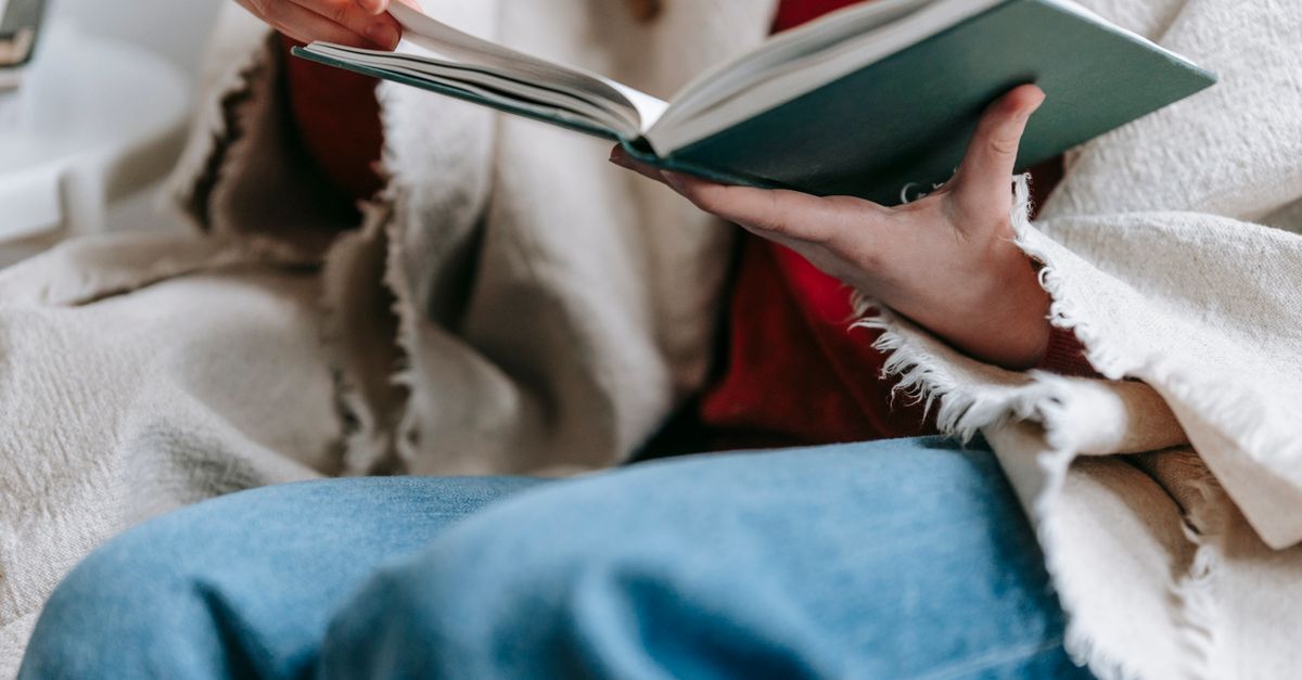 Crop anonymous lady in casual clothes and warm plaid on shoulders reading interesting book while sitting in armchair and spending free time at home